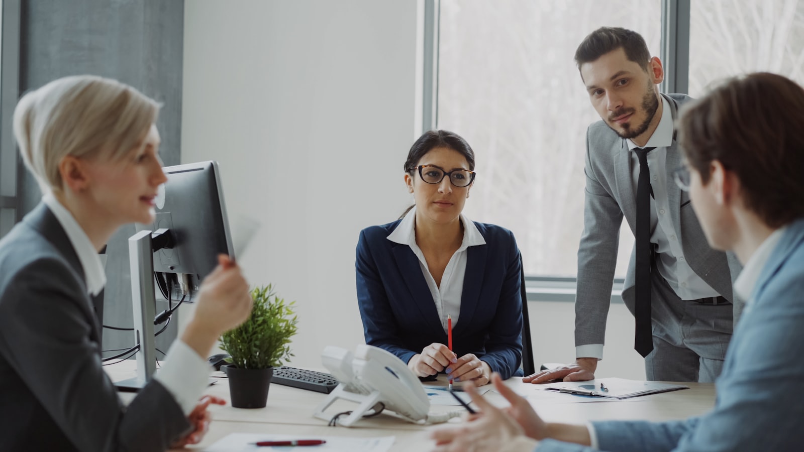 Four managers in business dress talking around a desk with a monitor and papers on it