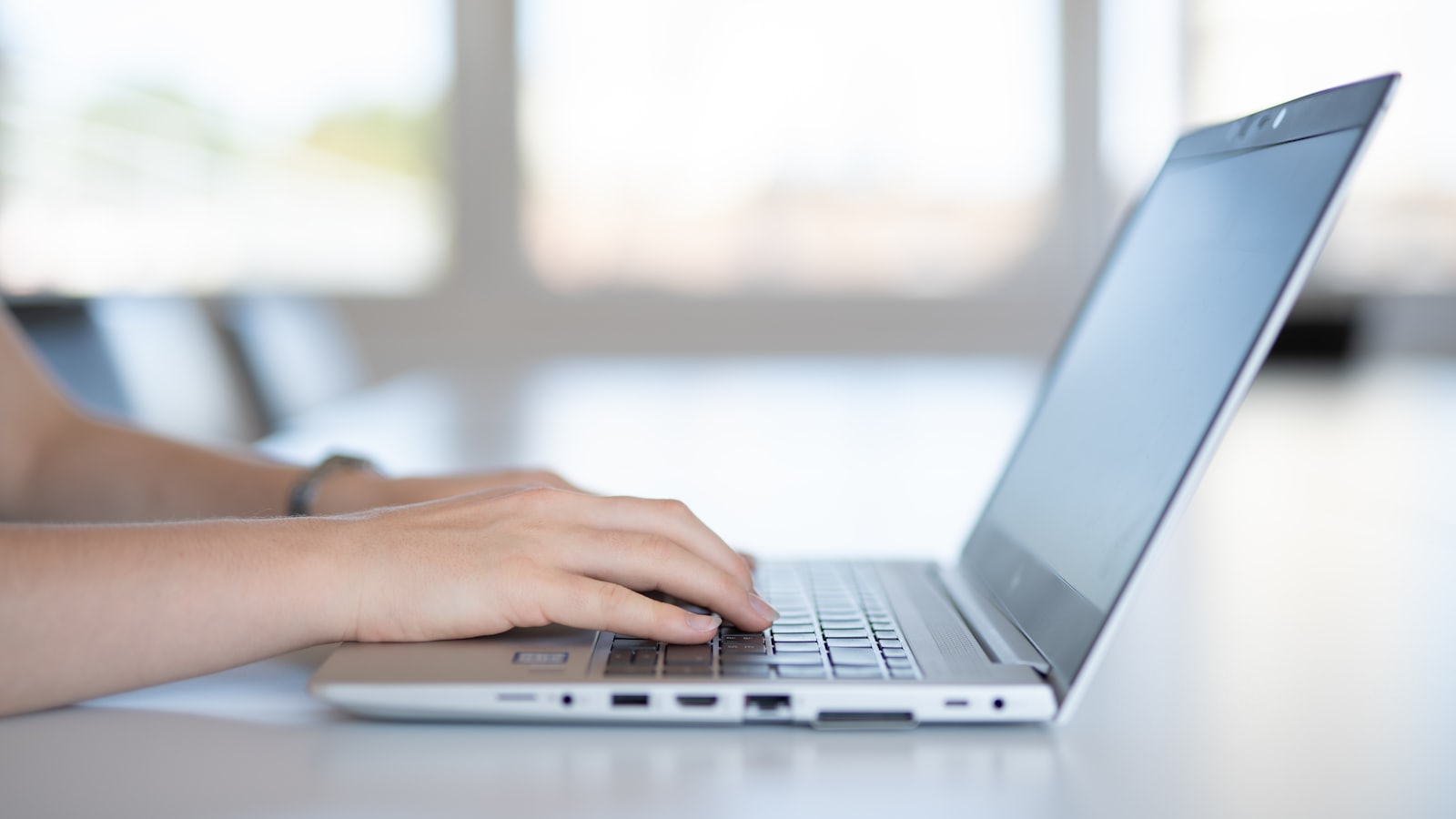 A hand typing on a laptop keyboard at a desk beside a bright window